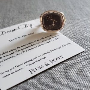 May include: A silver ring with a black wax seal featuring a telescope pointed at a constellation of stars. The ring is on a white card with the text "Dream Big" and "Look to the stars". The card also includes a quote by Vincent Van Gogh and the name "PLUM & POSEY".
