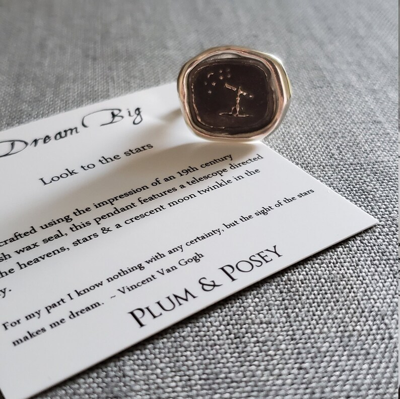 May include: A silver ring with a black wax seal featuring a telescope pointed at a constellation of stars. The ring is on a white card with the text "Dream Big" and "Look to the stars". The card also includes a quote by Vincent Van Gogh and the name "PLUM & POSEY".