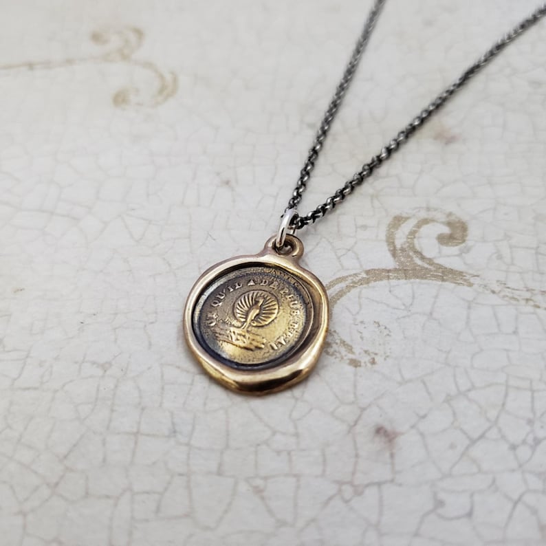 Puede incluir: Un collar dorado con un colgante redondo con un dise&ntilde;o de sello de cera. El colgante tiene una imagen en relieve de un p&aacute;jaro con las alas extendidas y el texto "De Quia Da Phusis" alrededor del borde.