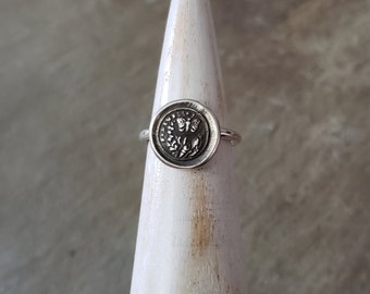 May include: A silver ring with a round, textured design featuring a butterfly and floral details.