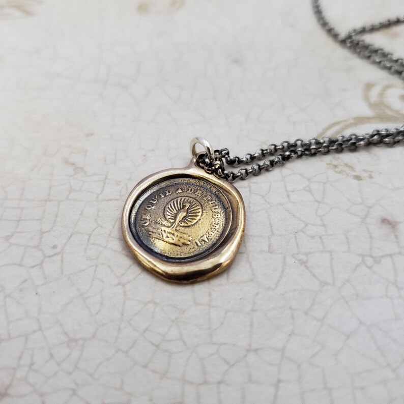 Puede incluir: Un collar con un colgante redondo de color dorado en forma de sello de cera, que presenta un dise&ntilde;o de p&aacute;jaro y el texto "Quod Vult Deus Adest Deus". El collar est&aacute; en una cadena plateada.