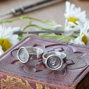 May include: Two silver rings with a deer head design on the top. The rings are sitting on a closed, red leather-bound book with gold leaf designs.
