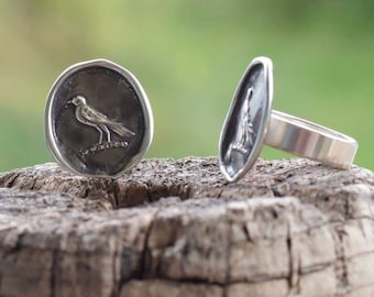 May include: A silver ring with a bird design on the face. The ring is shown from two angles, one with the bird design facing the camera and the other with the ring band facing the camera.