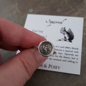 以下が含まれることがあります： リングの表面にリスのデザインが施された銀のリング。リングは指にはめられており、背景は「Squirrel」と「Ever Gay」の文字が書かれた白いカードです。
