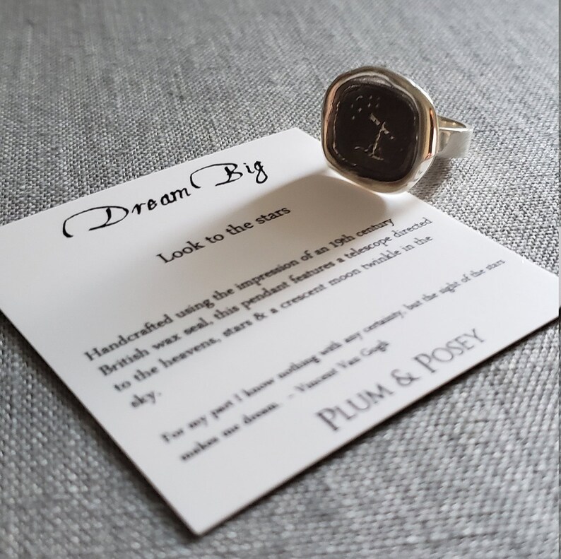 May include: A silver ring with a black wax seal featuring a telescope, stars, and a crescent moon. The ring is on a white card with the text "Dream Big" and "Look to the stars".