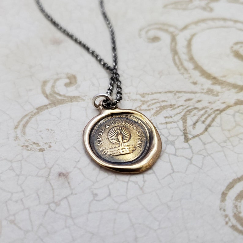 Puede incluir: Un collar con un colgante de sello de cera redondo con un dise&ntilde;o de f&eacute;nix y la inscripci&oacute;n "Qu'il a da plus beau". El collar est&aacute; en una cadena de plata.
