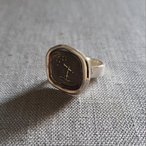 May include: A silver signet ring with a textured oval top featuring a stylized tree with stars above it.