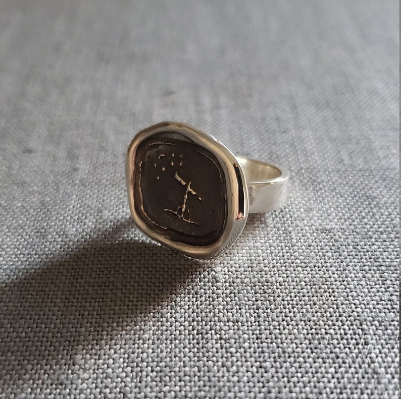 May include: A silver signet ring with a textured oval top featuring a stylized tree with stars above it.