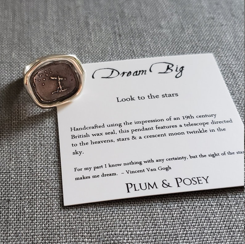 May include: Silver ring with a textured, octagonal shaped bezel featuring a vintage wax seal design with a telescope, stars, and a crescent moon. The ring is accompanied by a card with the text "Dream Big, Look to the stars" and a quote by Vincent Van Gogh. The card also includes the brand name "PLUM & POSEY".