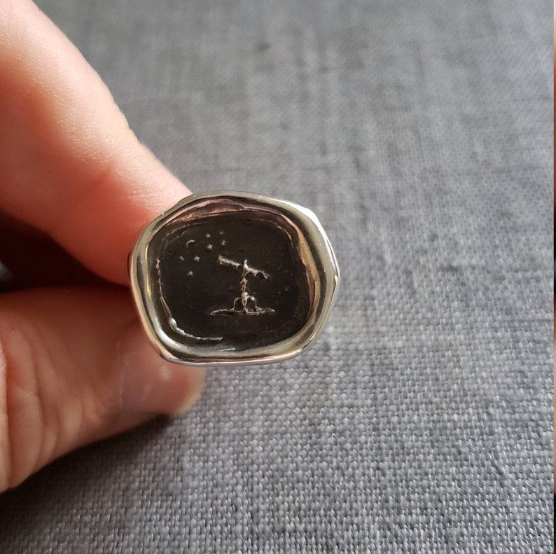 May include: A silver ring with a black wax seal featuring a small figure looking up at a constellation of stars.
