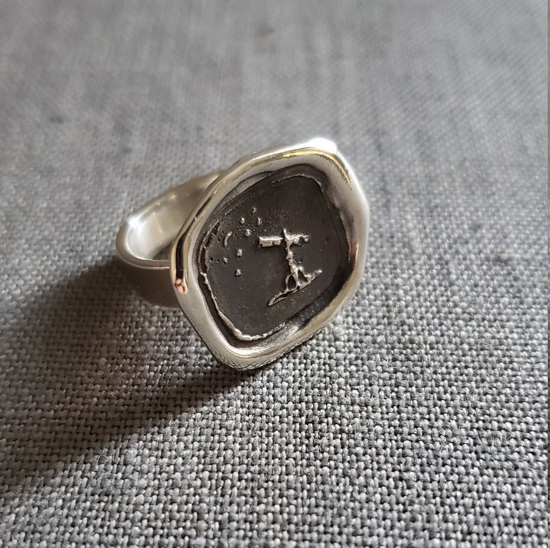 May include: A silver signet ring with a raised design of a cross and stars on a dark gray background.