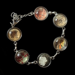May include: Silver-toned bracelet featuring seven circular charms with various designs. Charms include a butterfly, a flower, a portrait, and patterned paper. The bracelet has a chain closure and is set against a black background.