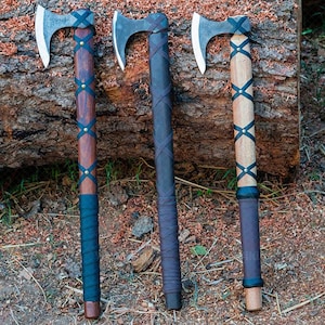 May include: Three axes with wooden handles and metal blades rest on a log. The handles are wrapped with leather and adorned with brass studs. The axe heads are silver, and the handles vary in colour from dark brown to light wood.