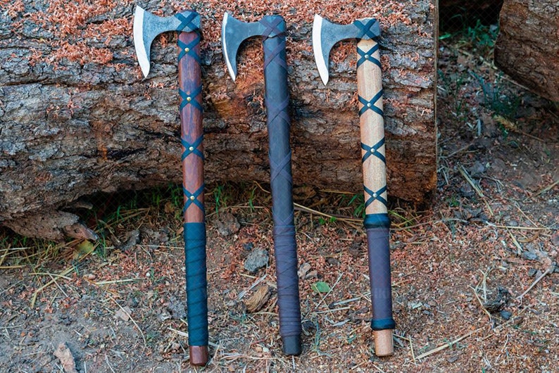 May include: Three axes with wooden handles and metal blades rest on a log. The handles are wrapped with leather and adorned with brass studs. The axe heads are silver, and the handles vary in color from dark brown to light wood.