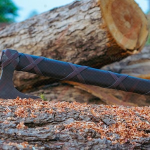 May include: A dark axe with a brown leather-wrapped handle and blade. The axe is resting on a wooden log covered in wood shavings. The axe's head is dark, and the handle is wrapped with crisscrossing leather straps.