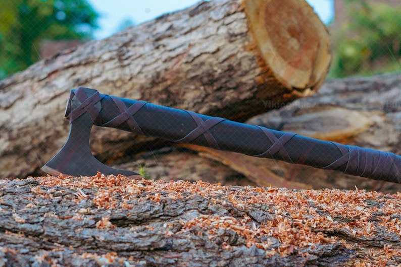 May include: A dark axe with a brown leather-wrapped handle and blade. The axe is resting on a wooden log covered in wood shavings. The axe's head is dark, and the handle is wrapped with crisscrossing leather straps.
