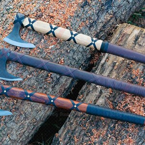 May include: Three axes with wooden handles and metal blades rest on a tree trunk. The handles are wrapped in leather and secured with black straps. The axe heads are a silver color, and the wood has a natural finish.