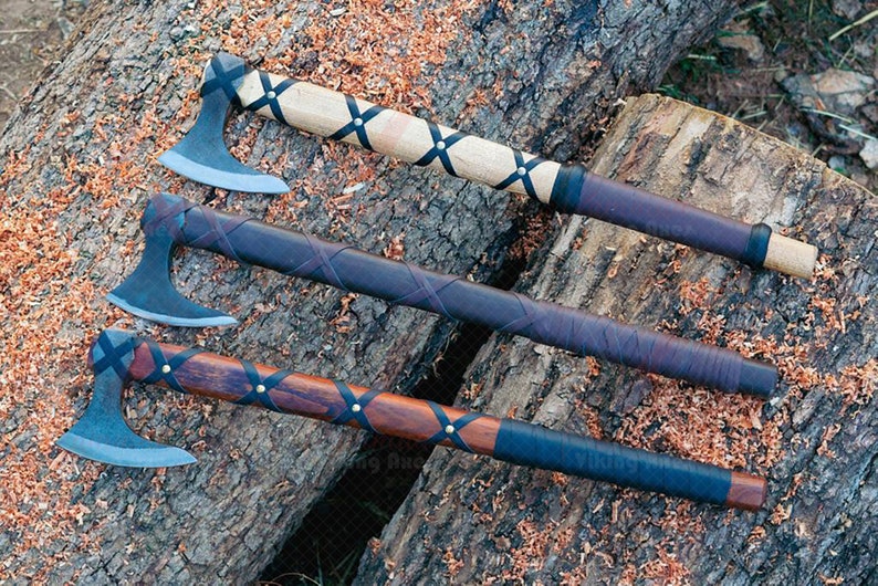 May include: Three axes with wooden handles and metal blades rest on a tree trunk. The handles are wrapped in leather and secured with black straps. The axe heads are a silver color, and the wood has a natural finish.