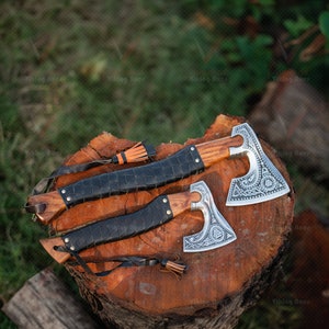 May include: Two Viking axes with leather wrapped handles and intricate metal designs on the blades. The axes are resting on a wooden stump.