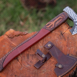Peut inclure: Une hache viking forgée à la main avec un manche en cuir et un fourreau en cuir. La tête de la hache est décorée de nœuds complexes et d'un motif celtique. Le manche est enveloppé de cuir brun et a un enroulement en cuir rouge à l'extrémité.