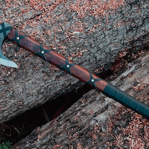May include: A handcrafted axe with a dark wooden handle and a steel blade. The handle is wrapped with black leather and secured with brass rivets. The axe is resting on a weathered log, with wood shavings scattered around.