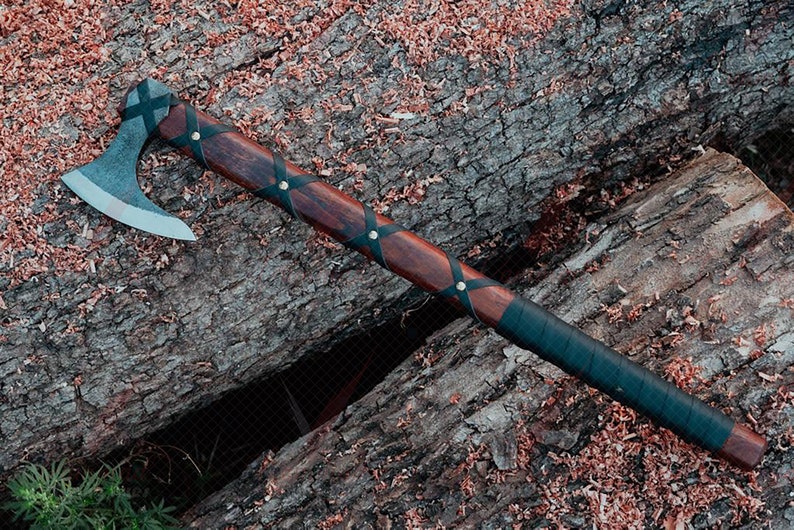 May include: A handcrafted axe with a dark wooden handle and a steel blade. The handle is wrapped with black leather and secured with brass rivets. The axe is resting on a weathered log, with wood shavings scattered around.