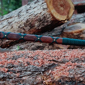May include: A handcrafted axe with a dark wooden handle wrapped in black leather and brass rivets. The axe head is made of metal. The axe is resting on a log with wood shavings.