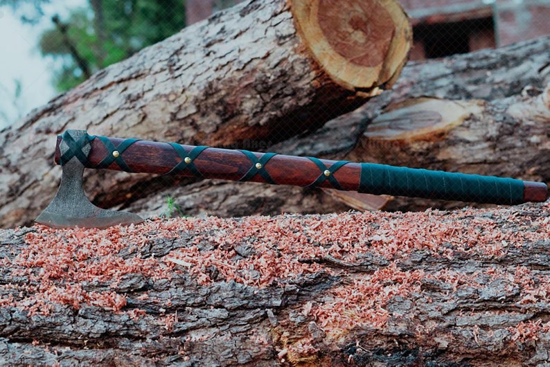 May include: A handcrafted axe with a dark wooden handle wrapped in black leather and brass rivets. The axe head is made of metal. The axe is resting on a log with wood shavings.