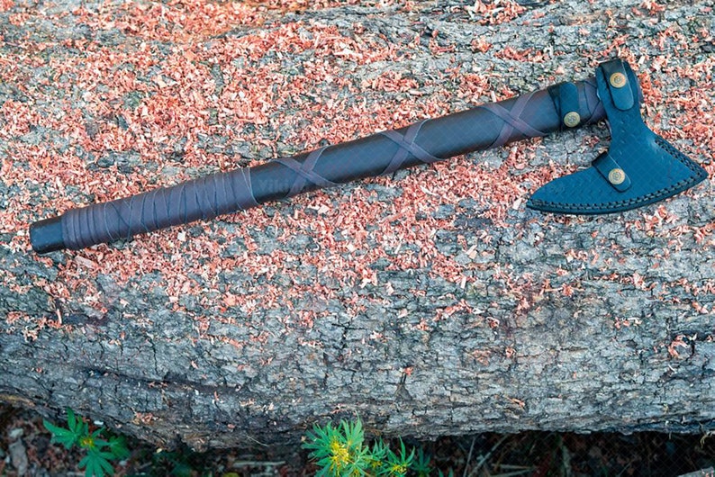 May include: A handcrafted axe with a dark brown handle wrapped in leather and a black axe head. The axe head has a leather sheath secured with brass-colored buttons. The axe is resting on a weathered log with wood shavings.