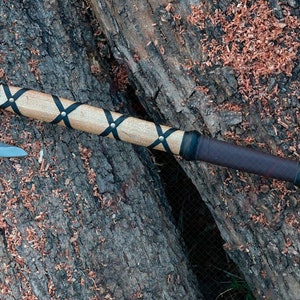 May include: A handcrafted axe with a gray steel blade and a light-colored wooden handle. The handle is wrapped with black leather straps and a dark brown leather grip. The axe rests on a textured wooden surface.