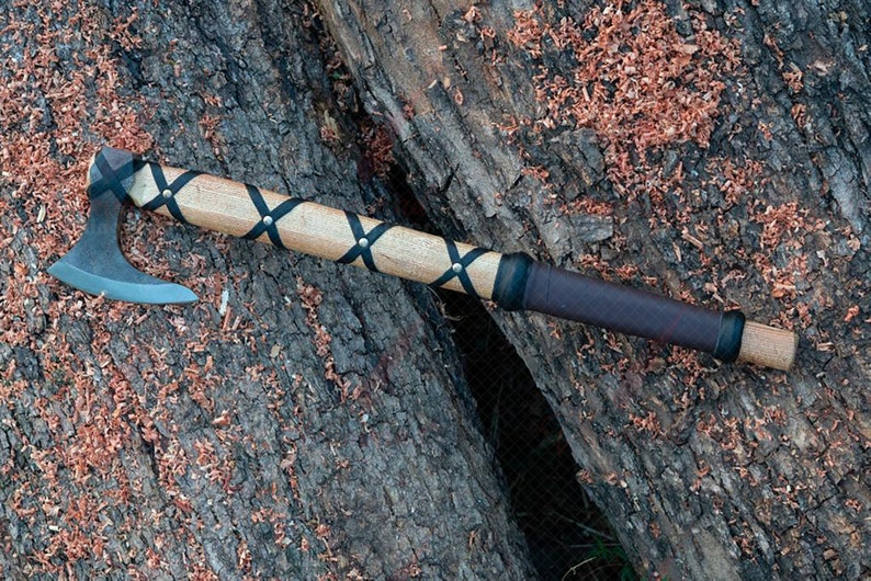 May include: A handcrafted axe with a gray steel blade and a light-colored wooden handle. The handle is wrapped with black leather straps and a dark brown leather grip. The axe rests on a textured wooden surface.
