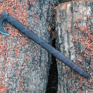 May include: A handcrafted axe with a dark wooden handle wrapped in leather. The axe head is made of metal. The axe is lying on a textured wooden surface with wood shavings.