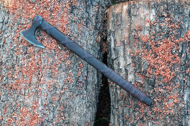 May include: A handcrafted axe with a dark wooden handle wrapped in leather. The axe head is made of metal. The axe is lying on a textured wooden surface with wood shavings.