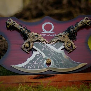 Può includere: Una targa in legno con sfondo rosso e le parole "God of War" in bianco. Due asce d'argento e oro sono montate sulla targa, con le lame incrociate. Le asce hanno disegni intricati e sono decorate con accenti dorati.