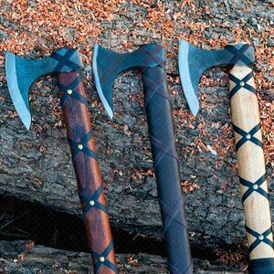 May include: Three Viking axes with steel blades and wooden handles. The handles are wrapped with black leather and secured with brass rivets. The axes rest on a weathered wooden surface with wood shavings.
