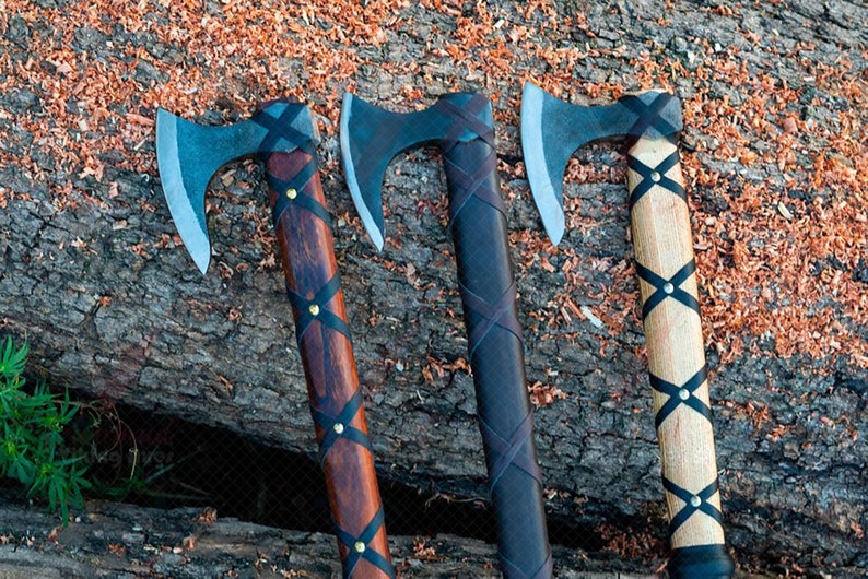 May include: Three Viking axes with steel blades and wooden handles. The handles are wrapped with black leather and secured with brass rivets. The axes rest on a weathered wooden surface with wood shavings.