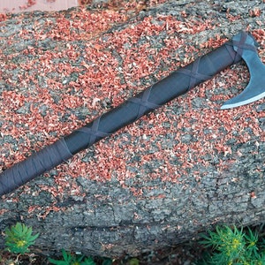 May include: A handcrafted axe with a dark handle wrapped in brown leather. The axe head is silver and the handle is wrapped in a crisscross pattern. The axe rests on a weathered log covered in wood shavings.