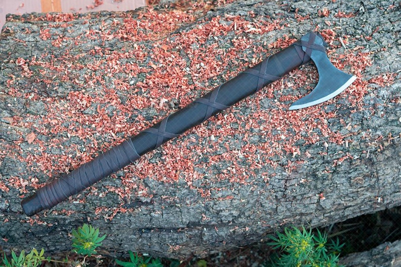 May include: A handcrafted axe with a dark handle wrapped in brown leather. The axe head is silver and the handle is wrapped in a crisscross pattern. The axe rests on a weathered log covered in wood shavings.