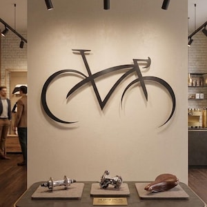 May include: A large black metal wall sculpture depicting a stylised bicycle silhouette mounted on a beige wall. Below, a table displays a silver bicycle hub, a silver derailleur, and a brown leather bicycle saddle with a small plaque.