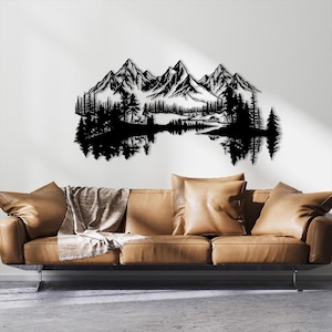 May include: Black metal wall art depicting a mountain range, trees, and a lake with reflections. The artwork is mounted on a white wall above a brown leather sofa with pillows and a blanket. The design evokes a nature theme.