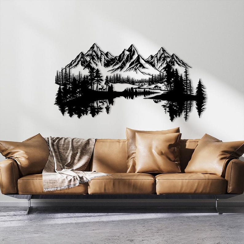 May include: Black metal wall art depicting a mountain range, trees, and a lake with reflections. The artwork is mounted on a white wall above a brown leather sofa with pillows and a blanket. The design evokes a nature theme.