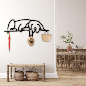 May include: Black metal wall-mounted coat rack shaped like a dog, with a red umbrella, a straw hat, and a straw bag hanging from it. The rack is mounted on a white wall, and there is a wooden bench with two wicker baskets underneath it.