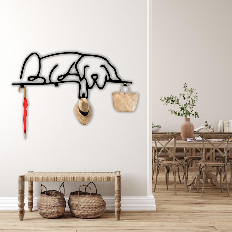 May include: Black metal wall-mounted coat rack shaped like a dog, with a red umbrella, a straw hat, and a straw bag hanging from it. The rack is mounted on a white wall, and there is a wooden bench with two wicker baskets underneath it.