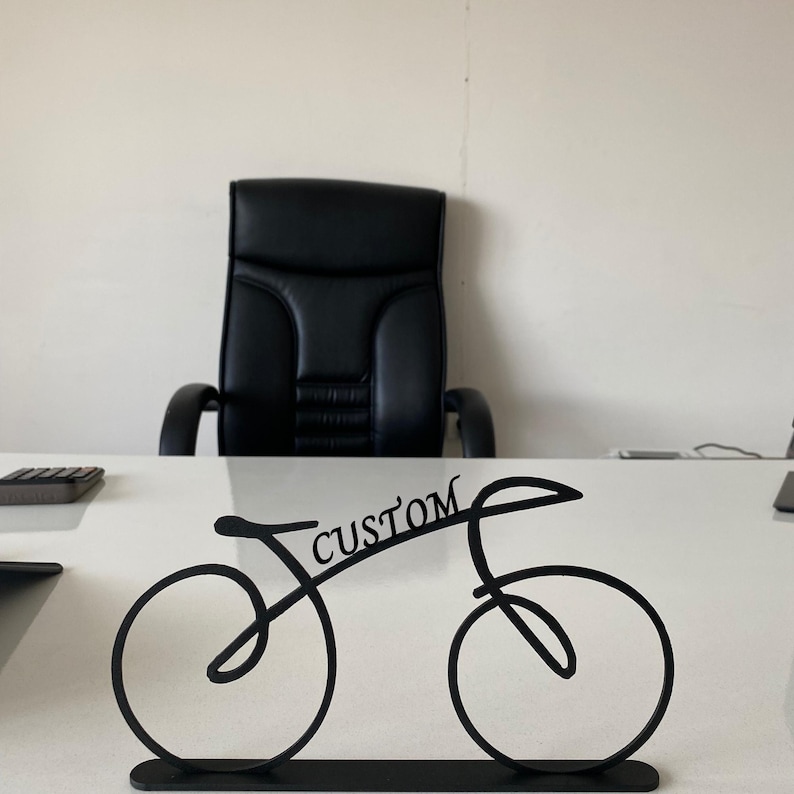 May include: Black metal bicycle-shaped desk tidy with the word "CUSTOM" in script. The bicycle design features two circular wheels and a curved frame. The tidy sits on a rectangular base and is placed on a white desk.