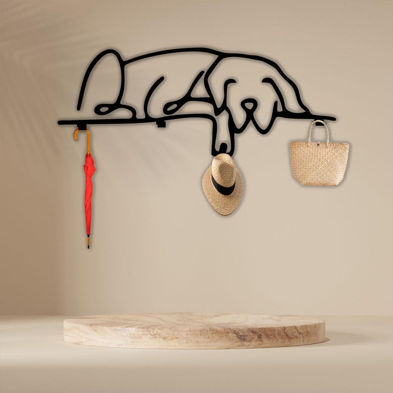 May include: Black metal wall hook shaped like a sleeping dog with a red umbrella, a straw hat, and a woven bag hanging from it.