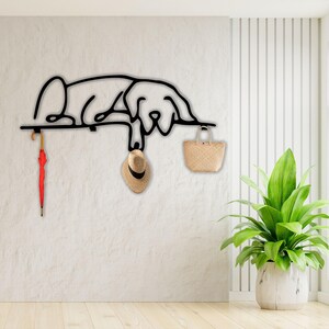May include: Black metal wall hook shaped like a dog lying down. The hook has a red umbrella, a straw hat, and a woven basket hanging from it.