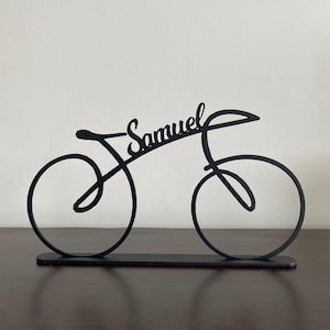 May include: Black metal bicycle sculpture with the name "Samuel" in cursive script. The bicycle is a minimalist design with two wheels, a seat, and handlebars, mounted on a rectangular base. The sculpture is displayed on a dark brown surface.