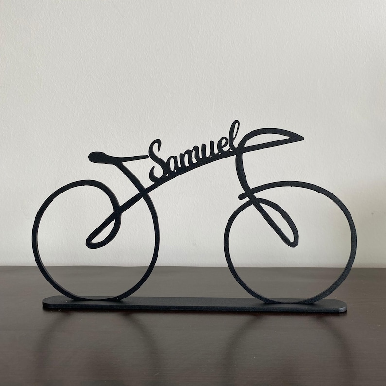 May include: Black metal bicycle sculpture with the name "Samuel" in cursive script. The bicycle is a minimalist design with two wheels, a seat, and handlebars, mounted on a rectangular base. The sculpture is displayed on a dark brown surface.