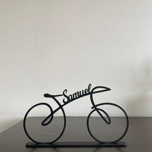 May include: A black metal bicycle sculpture with the name "Samuel" in cursive script. The bicycle design features two circular wheels, a seat, and handlebars. The sculpture is displayed on a dark surface against a white background.