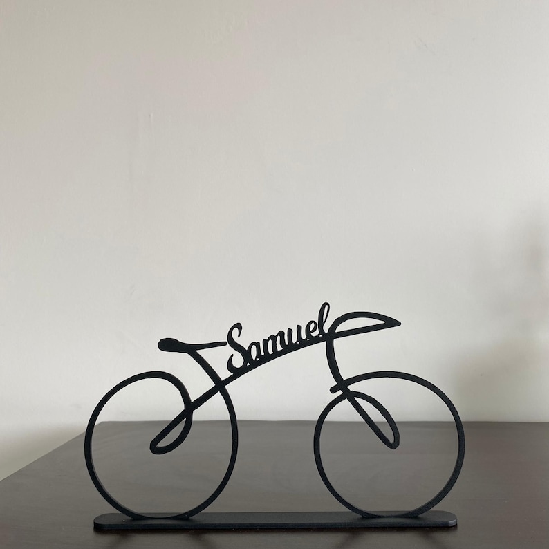 May include: A black metal bicycle sculpture with the name "Samuel" in cursive script. The bicycle design features two circular wheels, a seat, and handlebars. The sculpture is displayed on a dark surface against a white background.